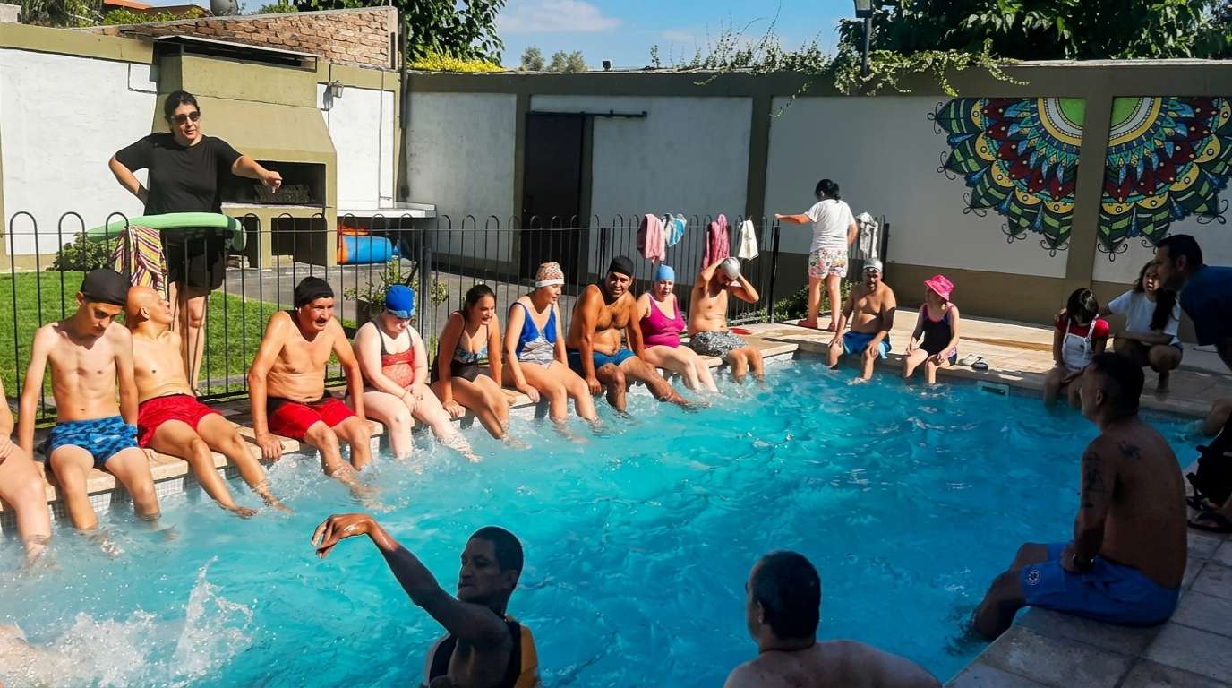 Pileta de natación con alumnos en traje de baño sentados en el borde y otros nadando, durante una actividad recreativa al aire libre.