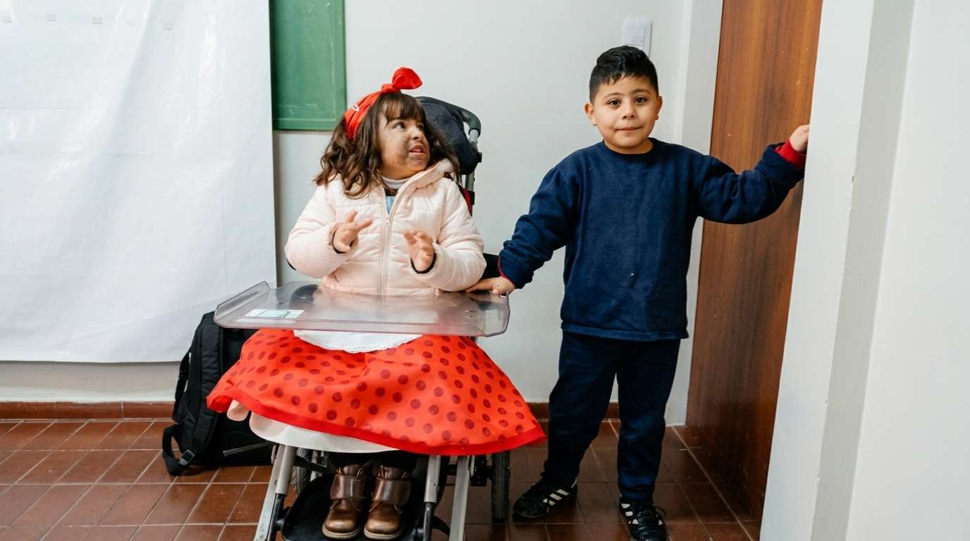 Una niña en silla de ruedas con vestido rojo a lunares y un compañero de la mano, en un pasillo del instituto.